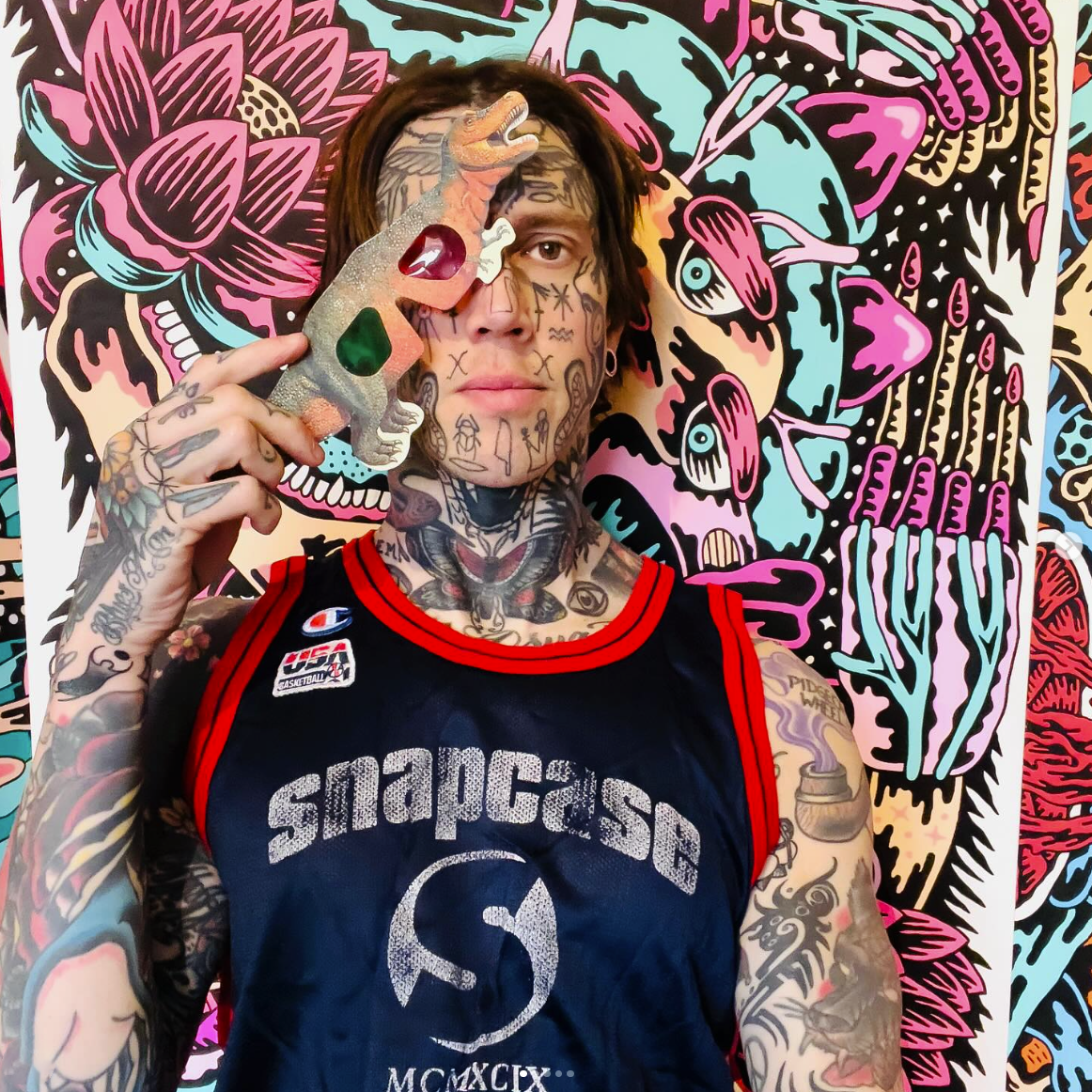 Marc UA, with extensive tattoos holds a pair of 3D-glasses against a background of his art work.
