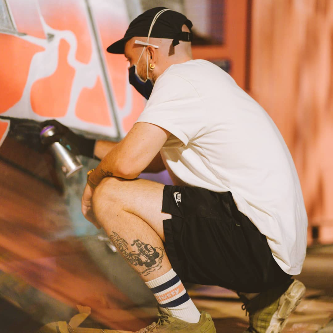 Bambi Bakbi in a white shirt and black shorts spray painting graffiti for a mural painting.