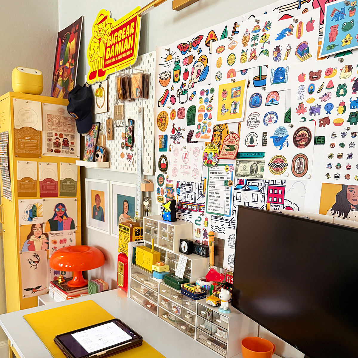 Colorful, eclectic workspace with a art-covered wall, desk accessories, a tablet, and a monitor. Bright, vibrant stickers.