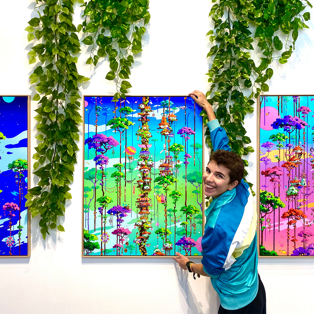 camila_illustration smiling, holding a colorful artwork of a vibrant, fantastical landscape. Green vines hang above them on the wall.