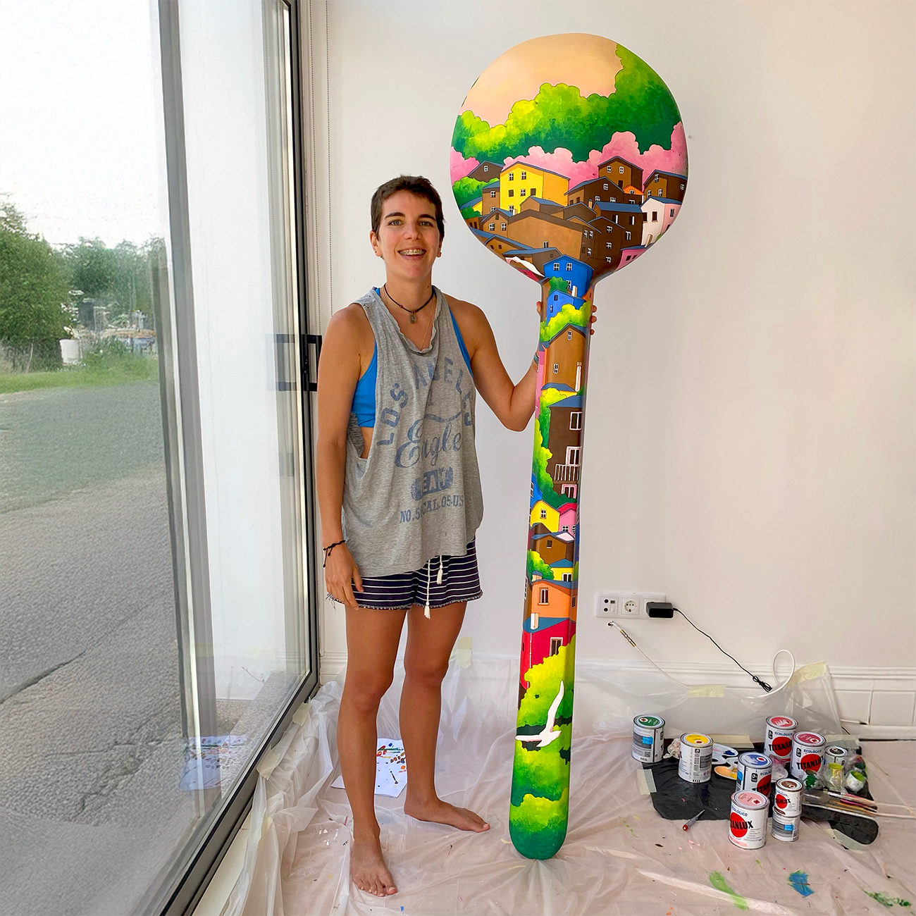 camila_illustration standing indoors, holding a large, colorful paintbrush with a painted landscape. Paint cans and brushes are on the floor nearby.