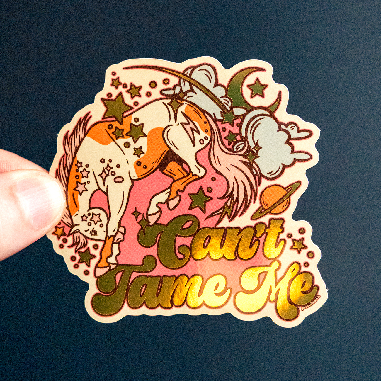 Gold mirror sticker of a colorful horde with stars and planets, accompanied by the text "Can't Tame Me," held by a hand against a dark background.