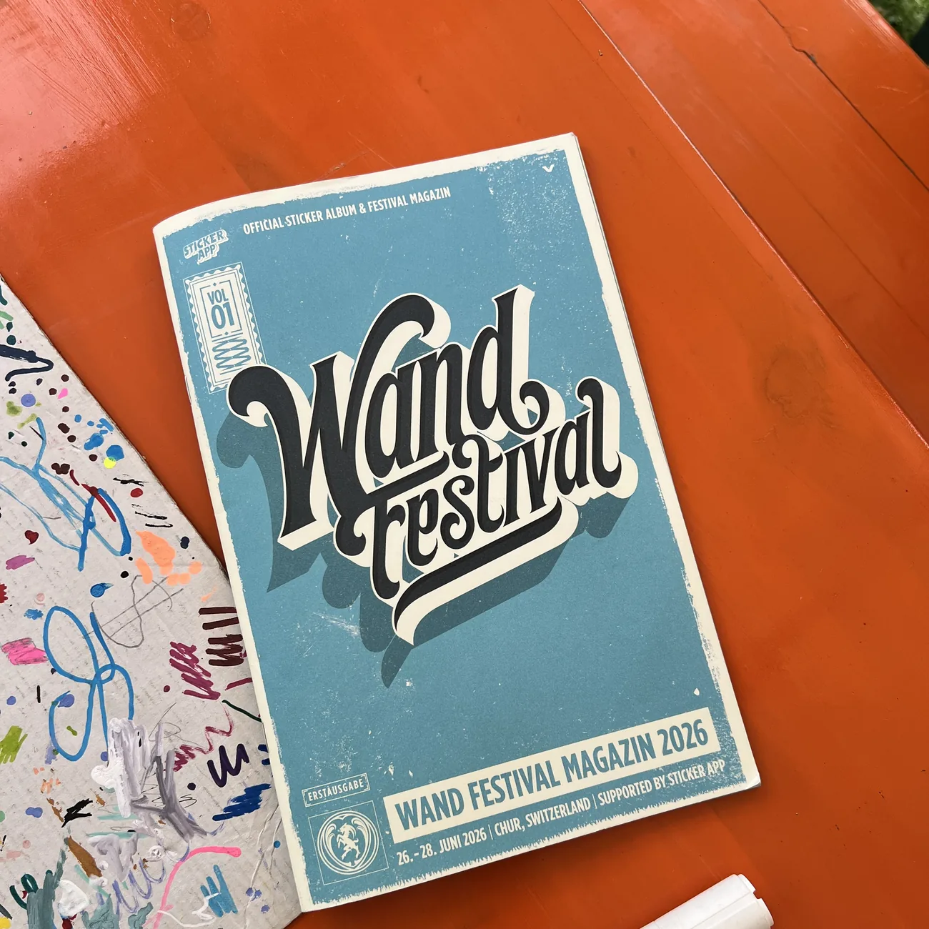 Offizielles Stickeralbum und Festivalmagazin des Wand Festival 2026, unterstützt von StickerApp, neben einem Paint-Marker auf einem orangefarbenen Tisch