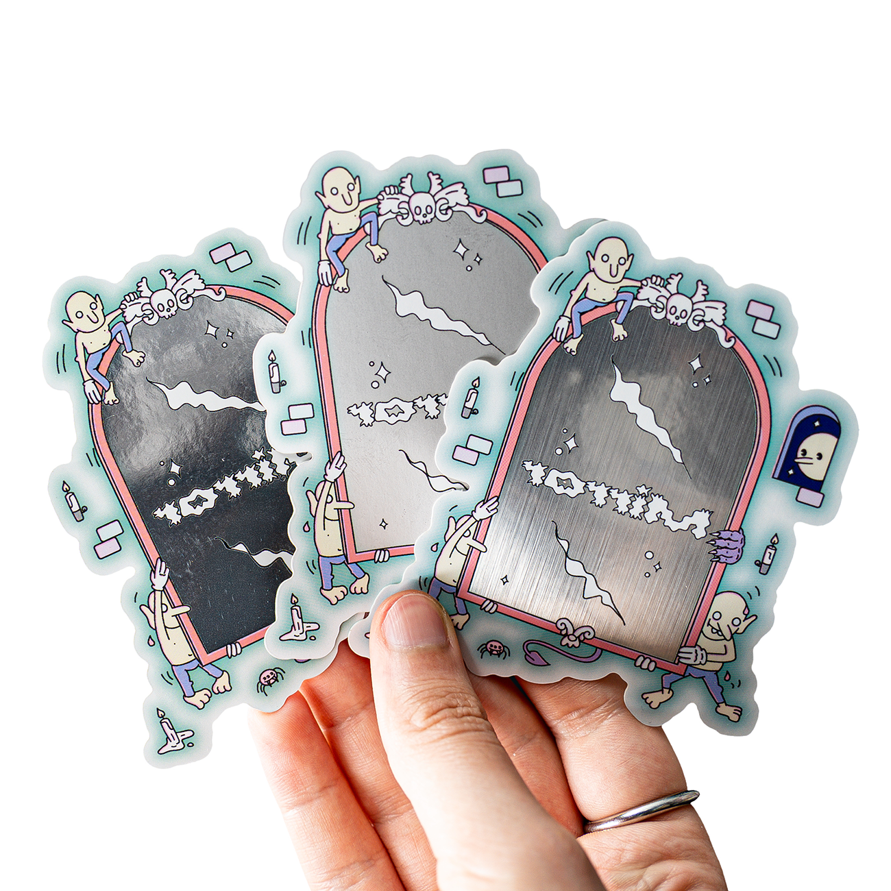 Hand holding three metallic stickers featuring cartoon skeletons climbing on silver mirrors with playful designs.