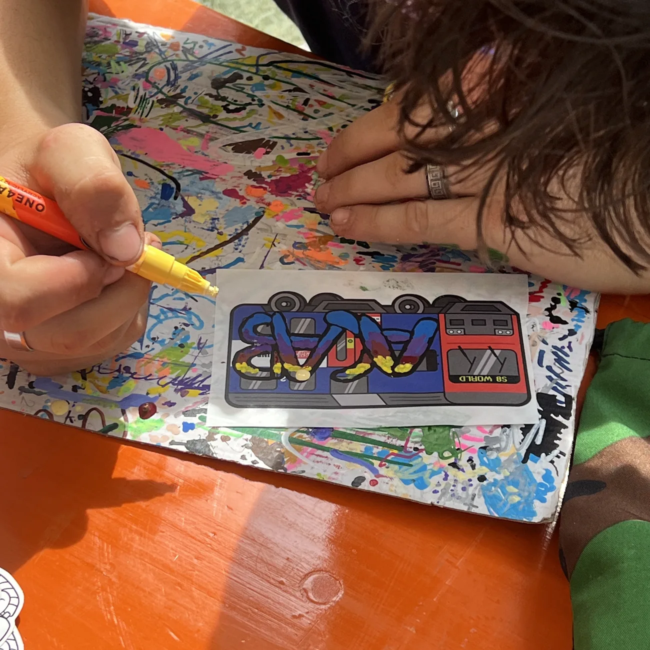 Eine Hand malt ein Etikett mit dem S8-World-Zug mit einem gelben Paint-Marker aus, beim Workshop von Brainfart x StickerApp