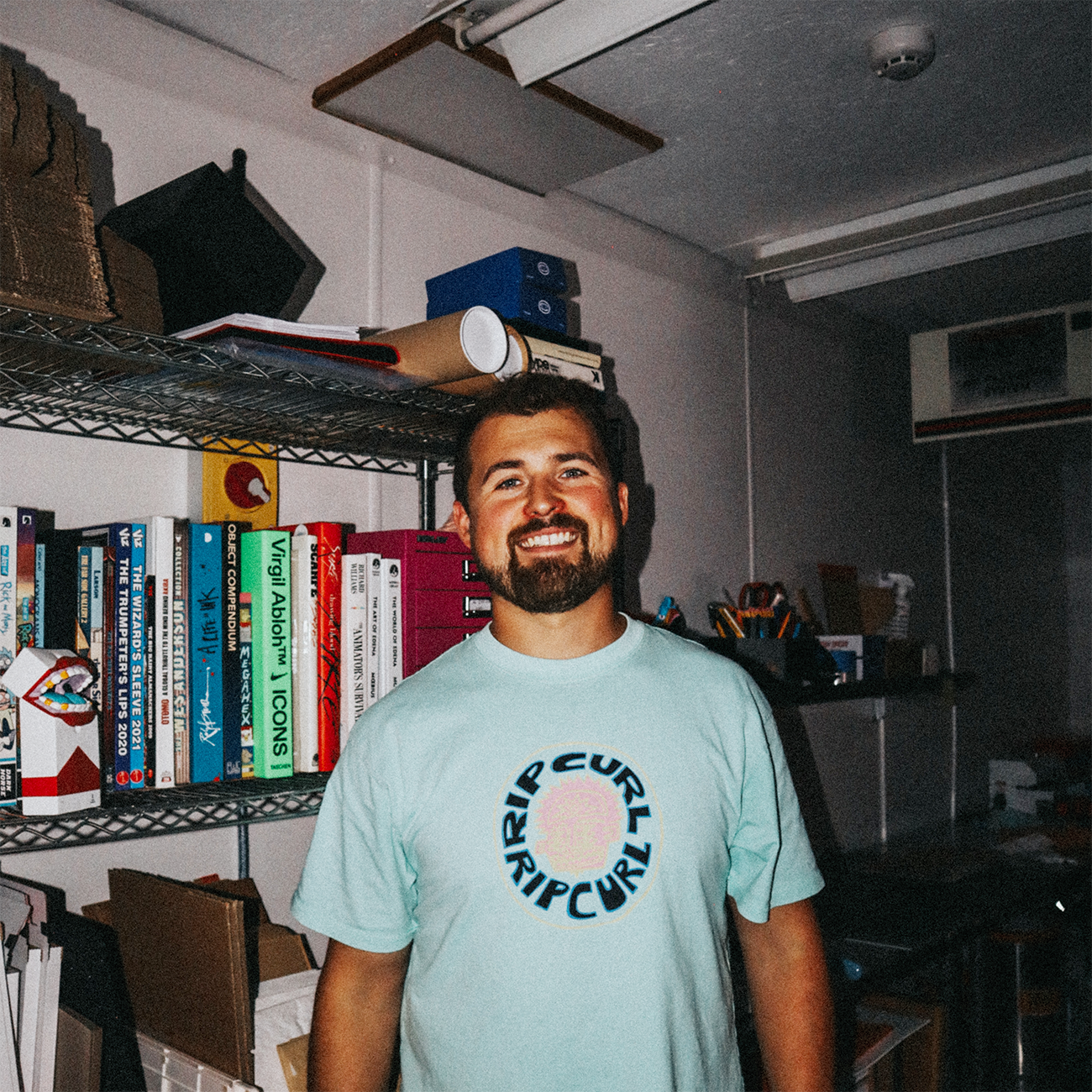Sam Drew, aka Lumps, smiling in a room with shelves of books and office supplies.