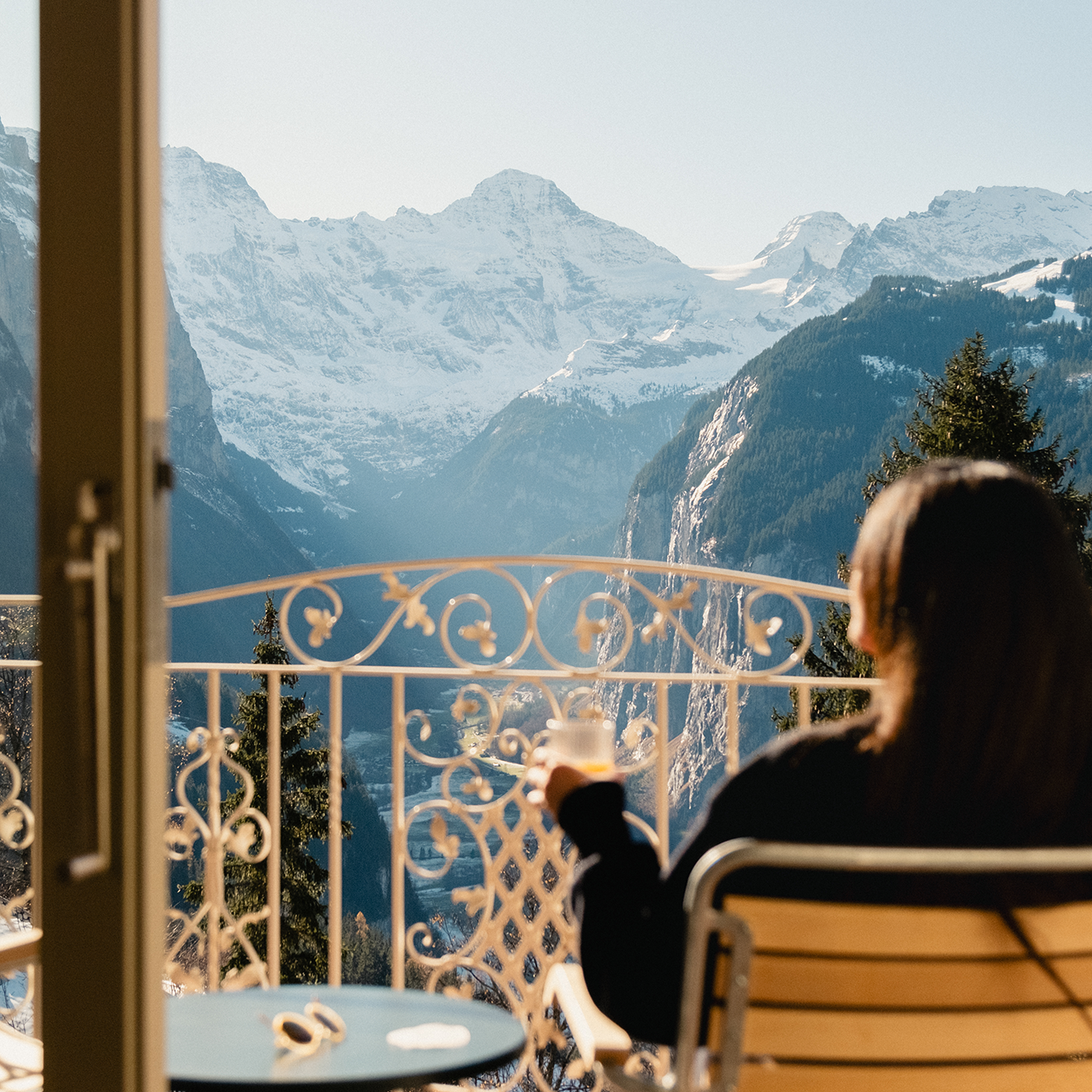 Person sitting on a balcony with a drink, overlooking snow-capped mountains and a clear blue sky.