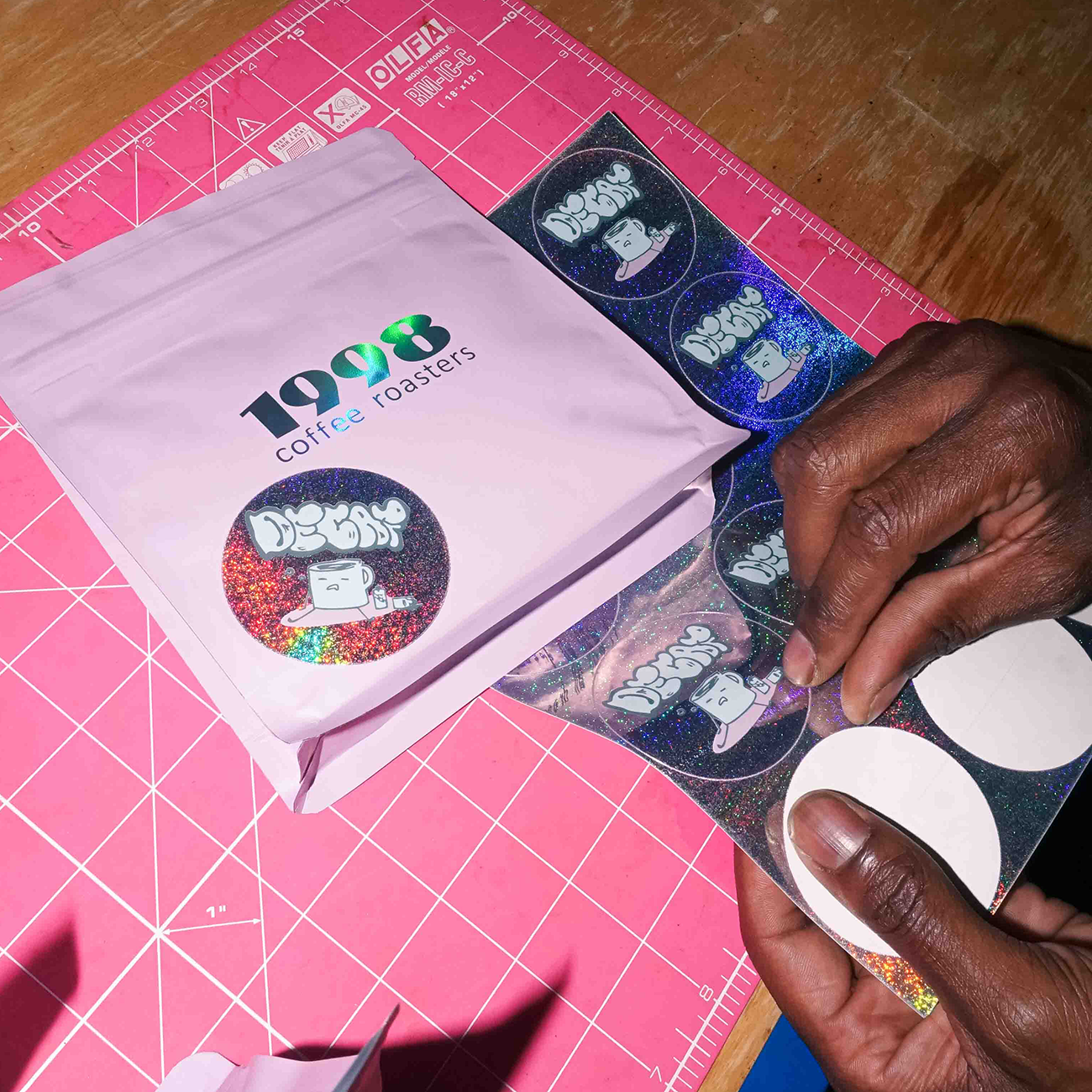 Hands applying Pixie Dust stickers to a pink coffee bag labeled "1908 coffee roasters" on a pink cutting mat.