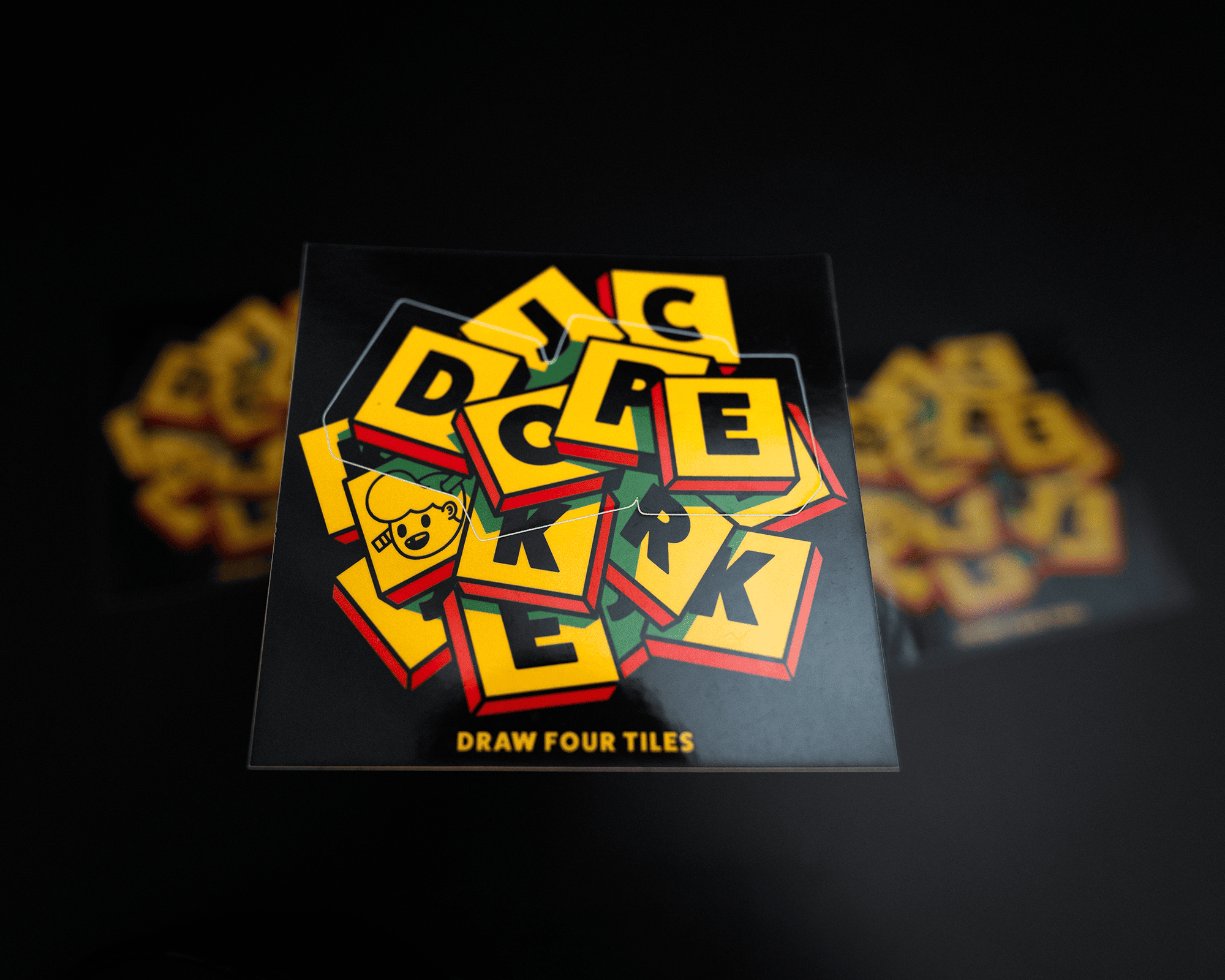 A kiss cut sticker showing a stack of colorful tiles with letters and symbols, including a clover and smiley face, on a black background. Text reads "Draw Four Tiles."