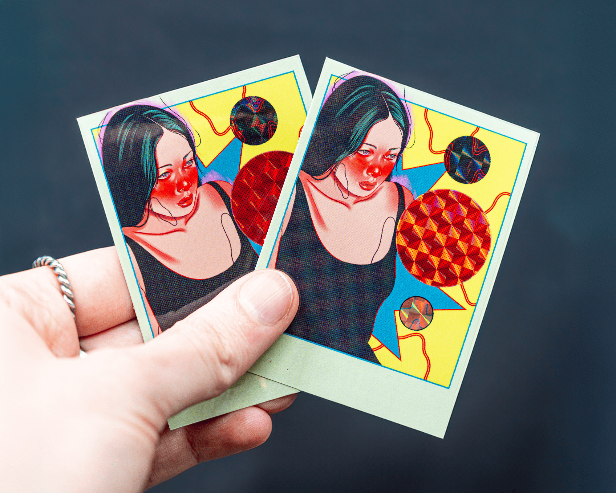 Hand holding two vibrant, artistic prismatic stickers featuring a woman with red cheeks, colorful geometric shapes, and a dark background.