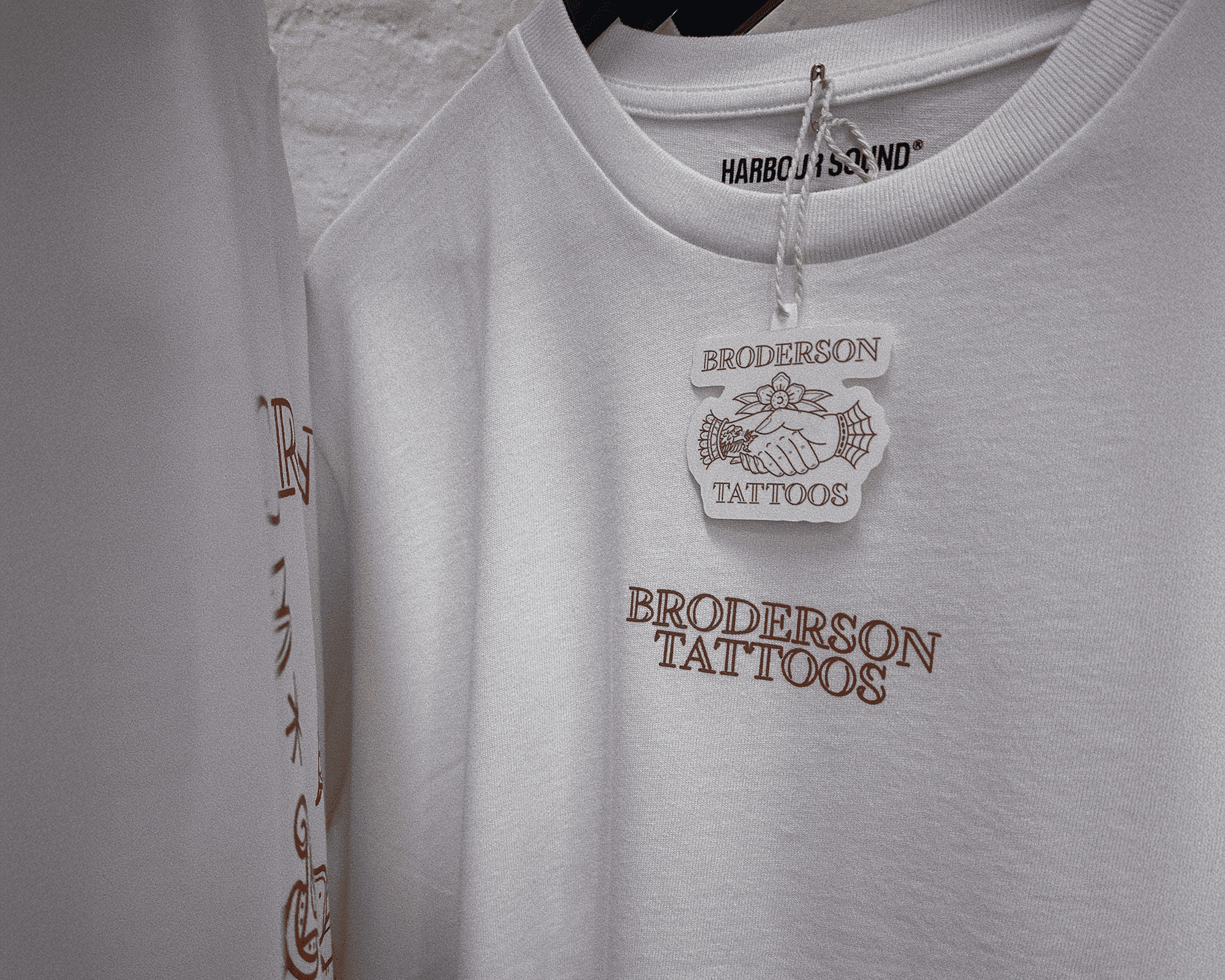 White T-shirt with "Brodersøn Tattoos" printed in brown, hanging on a rack with a branded hang tag attached.