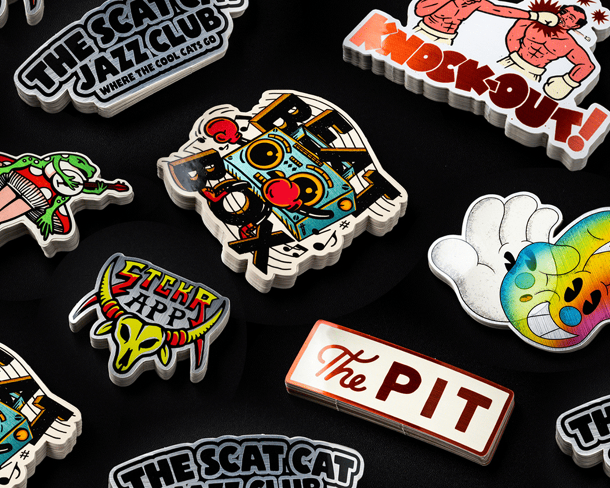 A collection of colorful, diverse brushed aluminum stickers on a black background, featuring various designs and text like "The Pit" and "Red Box."