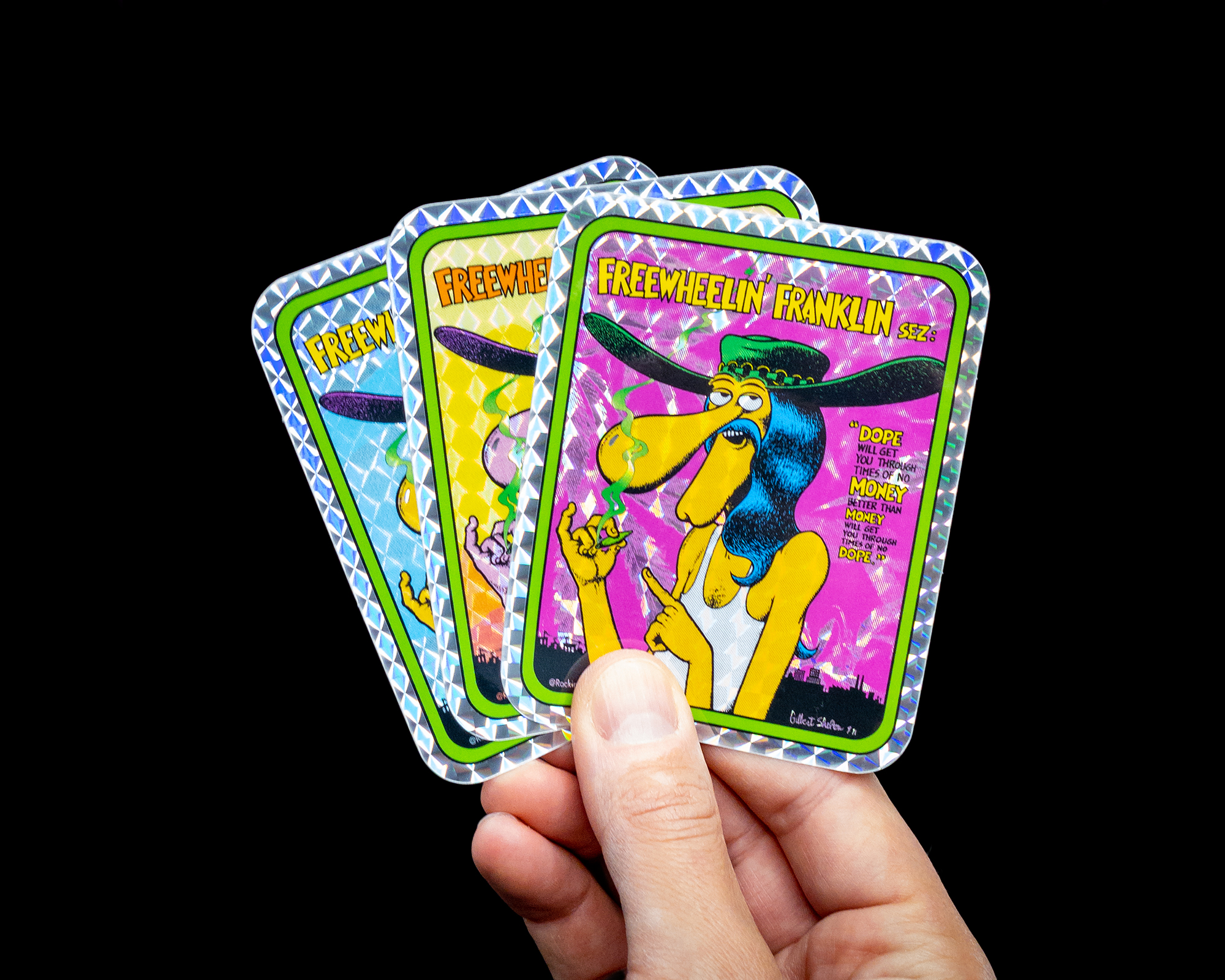 Hand holding four prismatic "Freewheelin' Franklin" sticker cards with psychedelic artwork and a cartoon character on a black background.