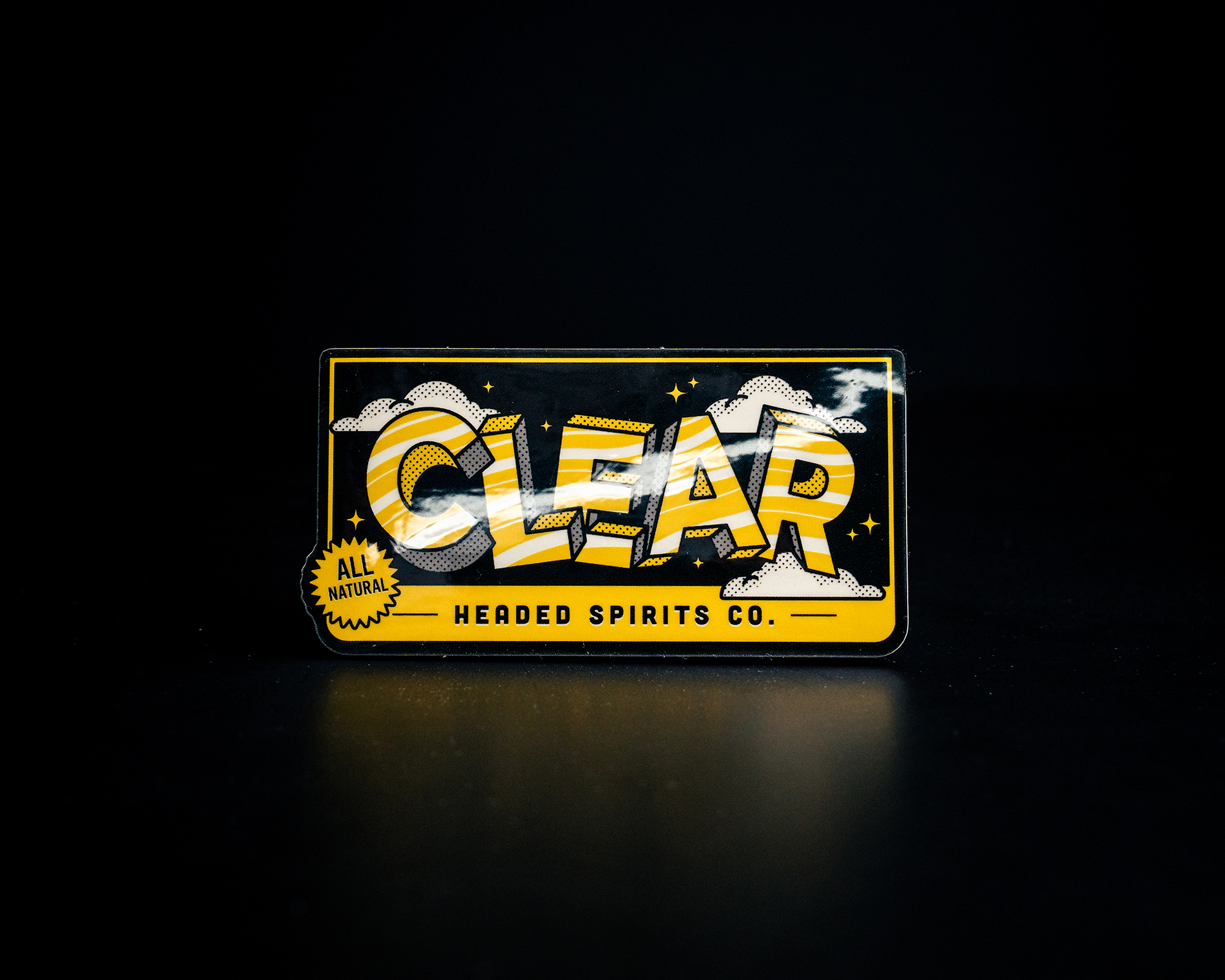 Close-up of a colorful "CLEAR Headed Spirits Co." sticker with a bold design and all-natural label, set against a dark background.