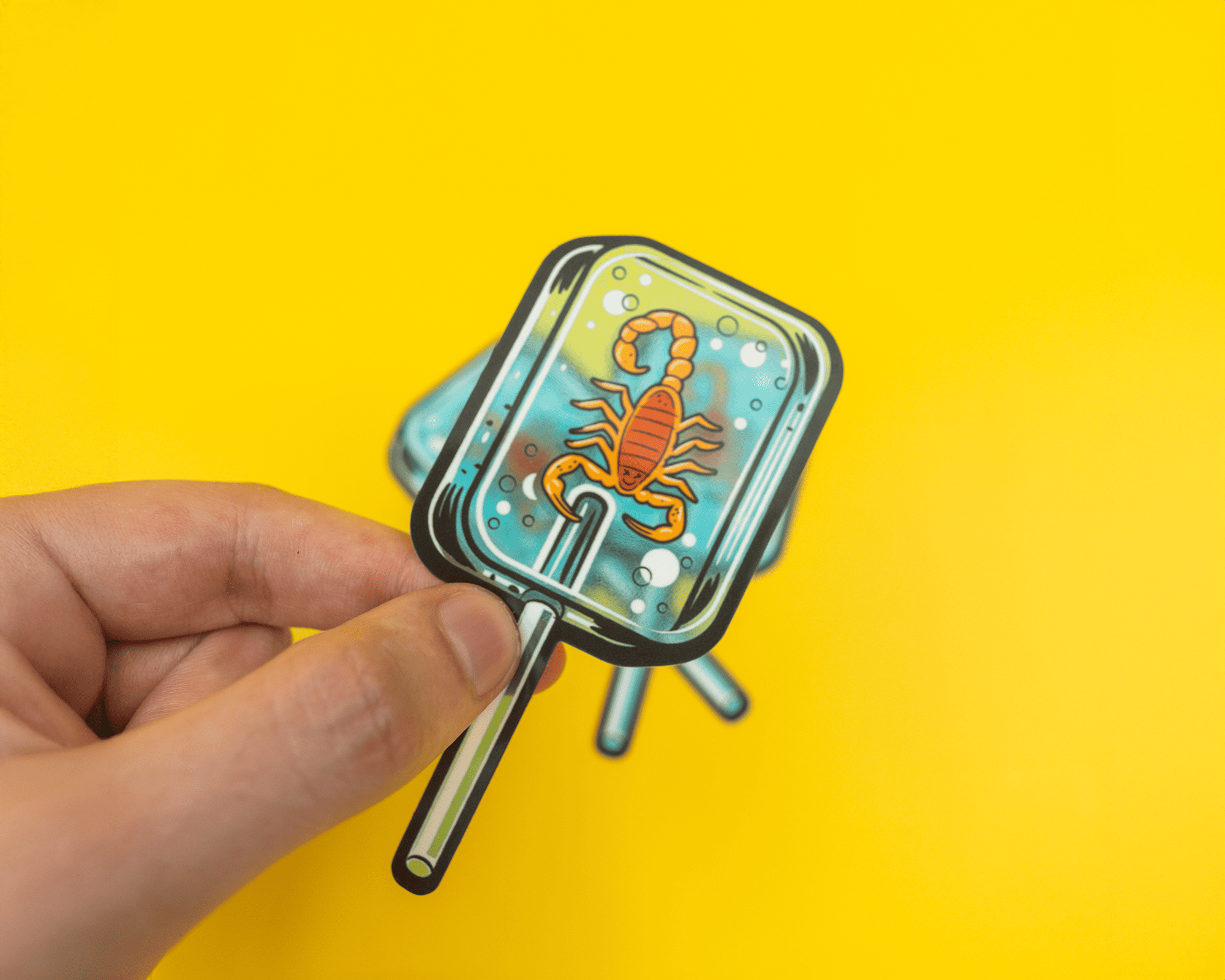 Hand holding a transparent lollipop sticker with a scorpion inside against a bright yellow background.