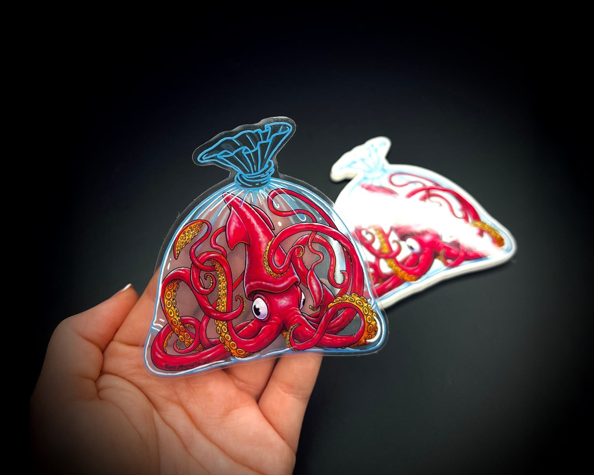 A hand holds a transparent sticker of a red octopus in a plastic bag design. Another similar sticker is in the background on a dark surface.