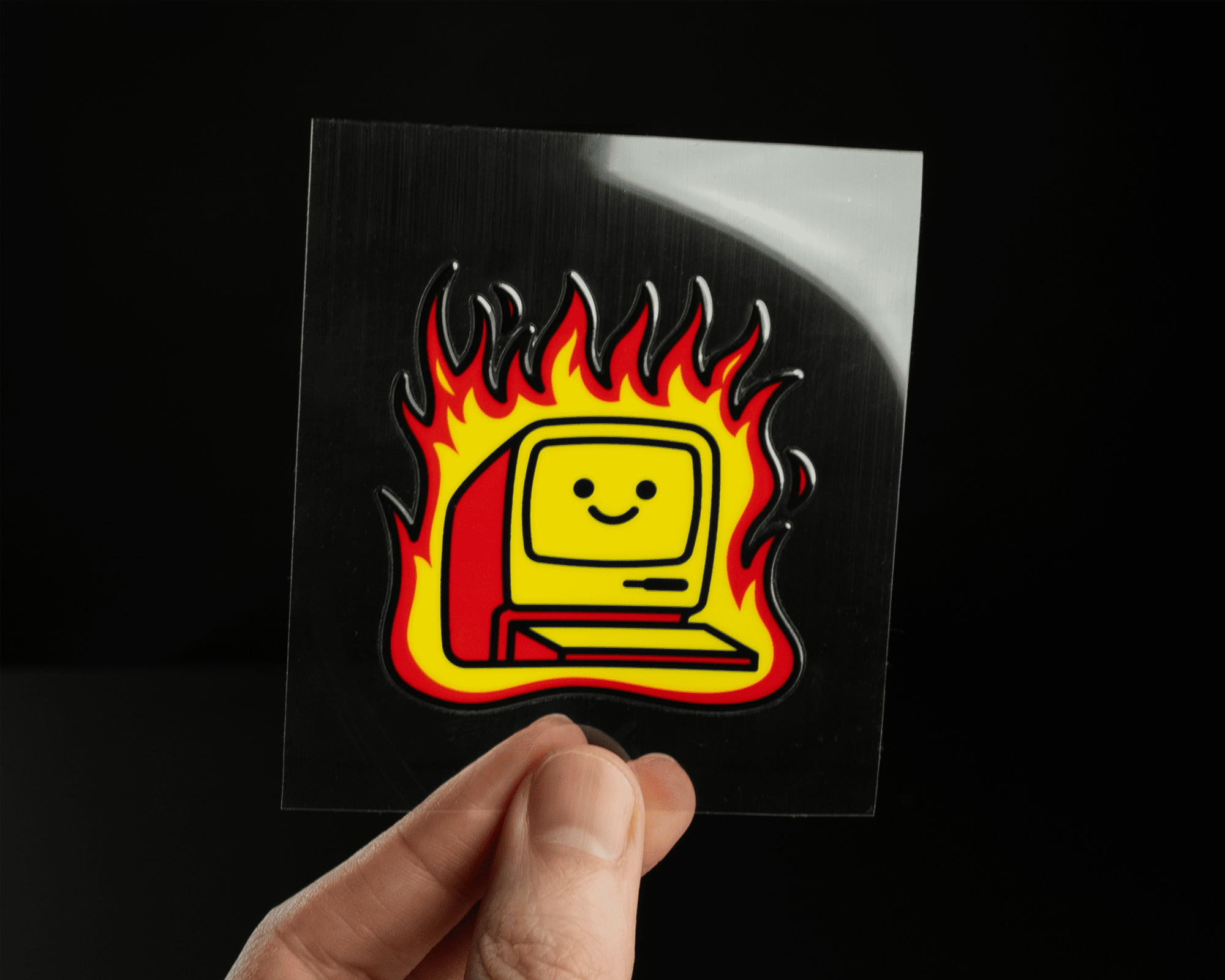Hand holding a UV-Dtf sticker of a smiling vintage computer with flames around it, against a dark background.