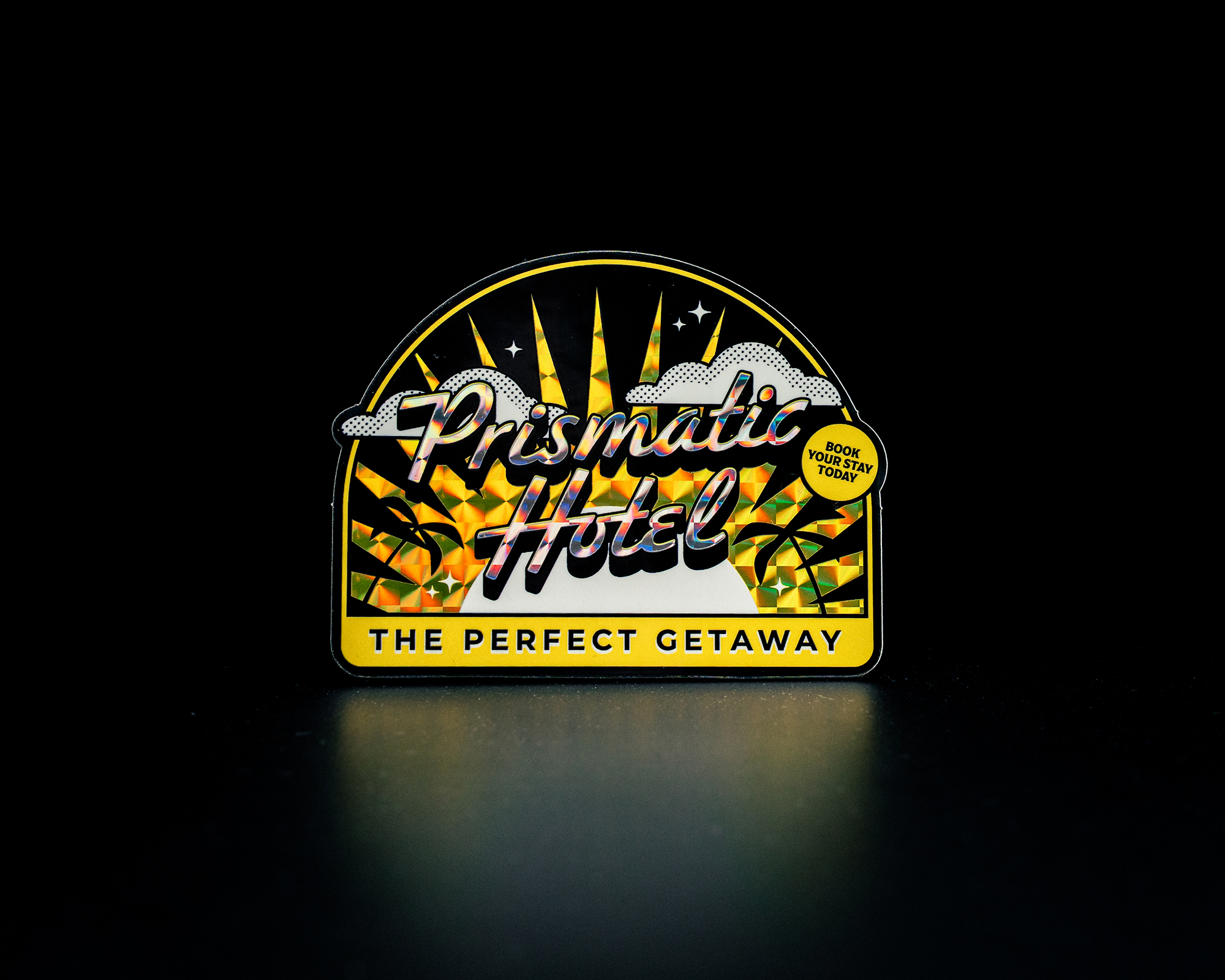 Sticker of Retro-style "Prismatic Hotel" sign with sunburst, clouds, and palm trees on a black background; reads "The Perfect Getaway" and "Book Your Stay Today."