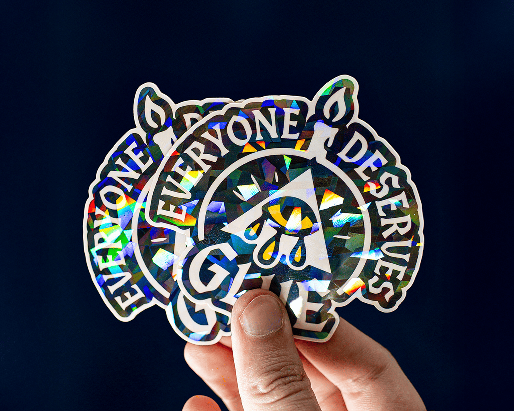Hand holding holographic cracked ice stickers with the text "Everyone Deserves Glue" and a design featuring a candle and a triangle with a droplet.