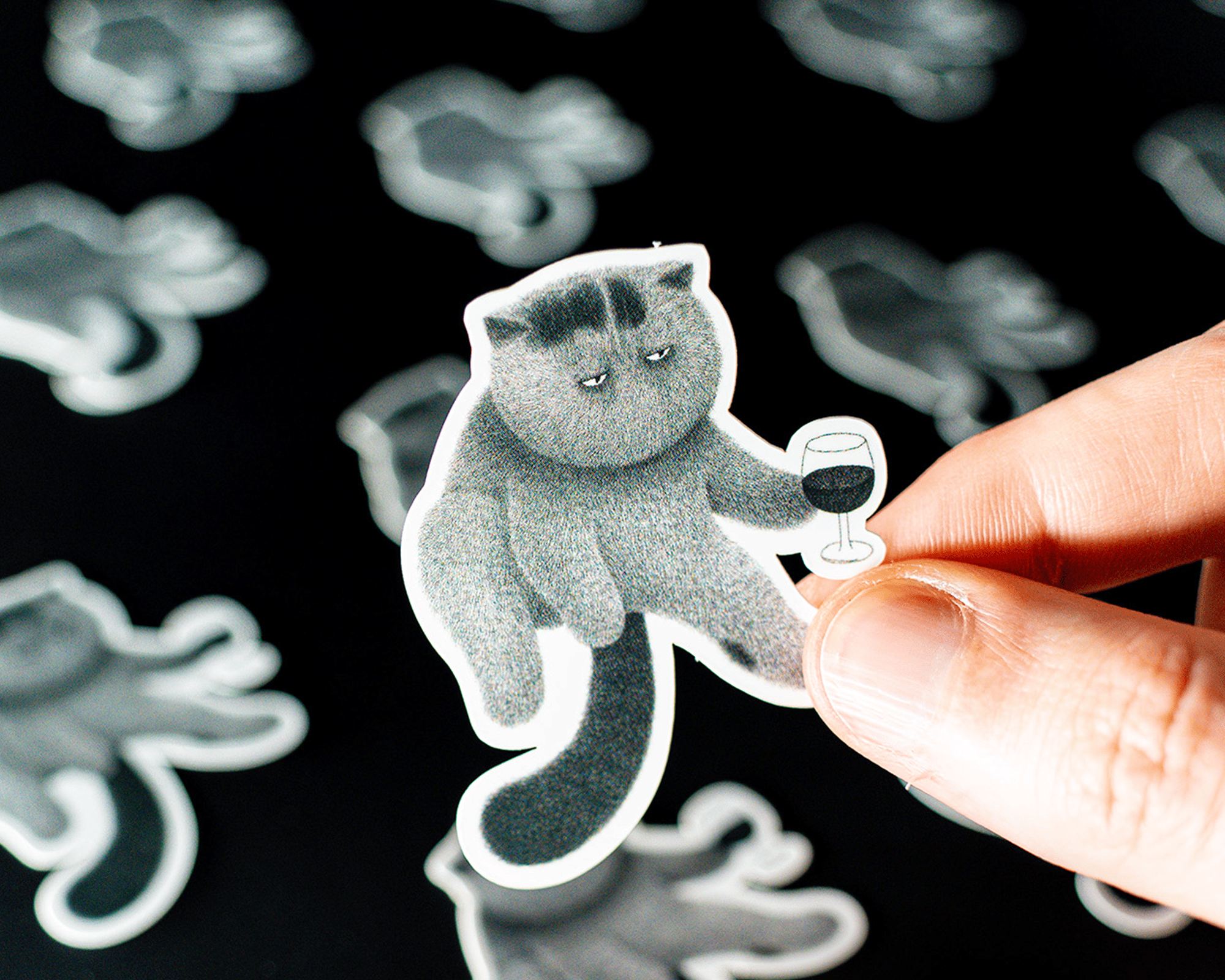 Vinyl sticker of a grumpy cat holding a wine glass, with several similar stickers blurred in the background.