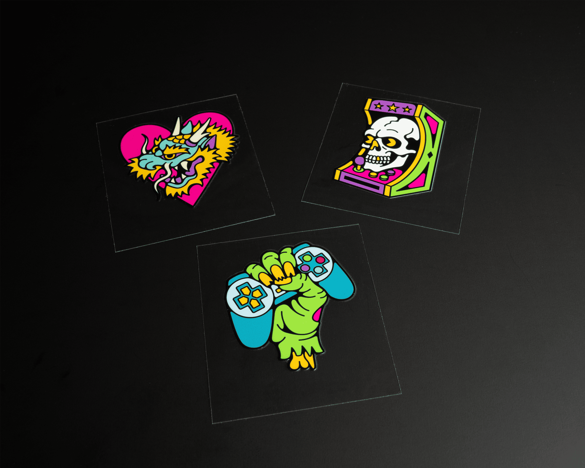 Three colorful uv dtf stickers on a black background: a dragon in a heart, a skull in an arcade machine, and a green hand holding a game controller.