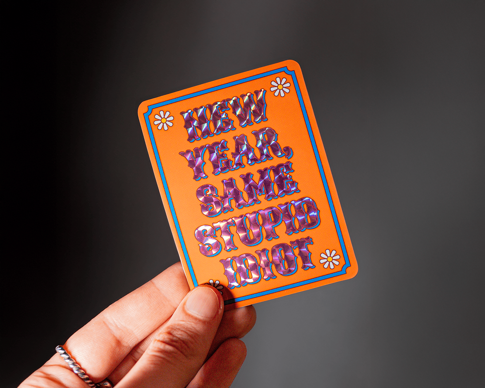 Hand holding an orange prismatic sticker with colorful text saying, "NEW YEAR, SAME STUPID IDIOT," adorned with small flower designs.