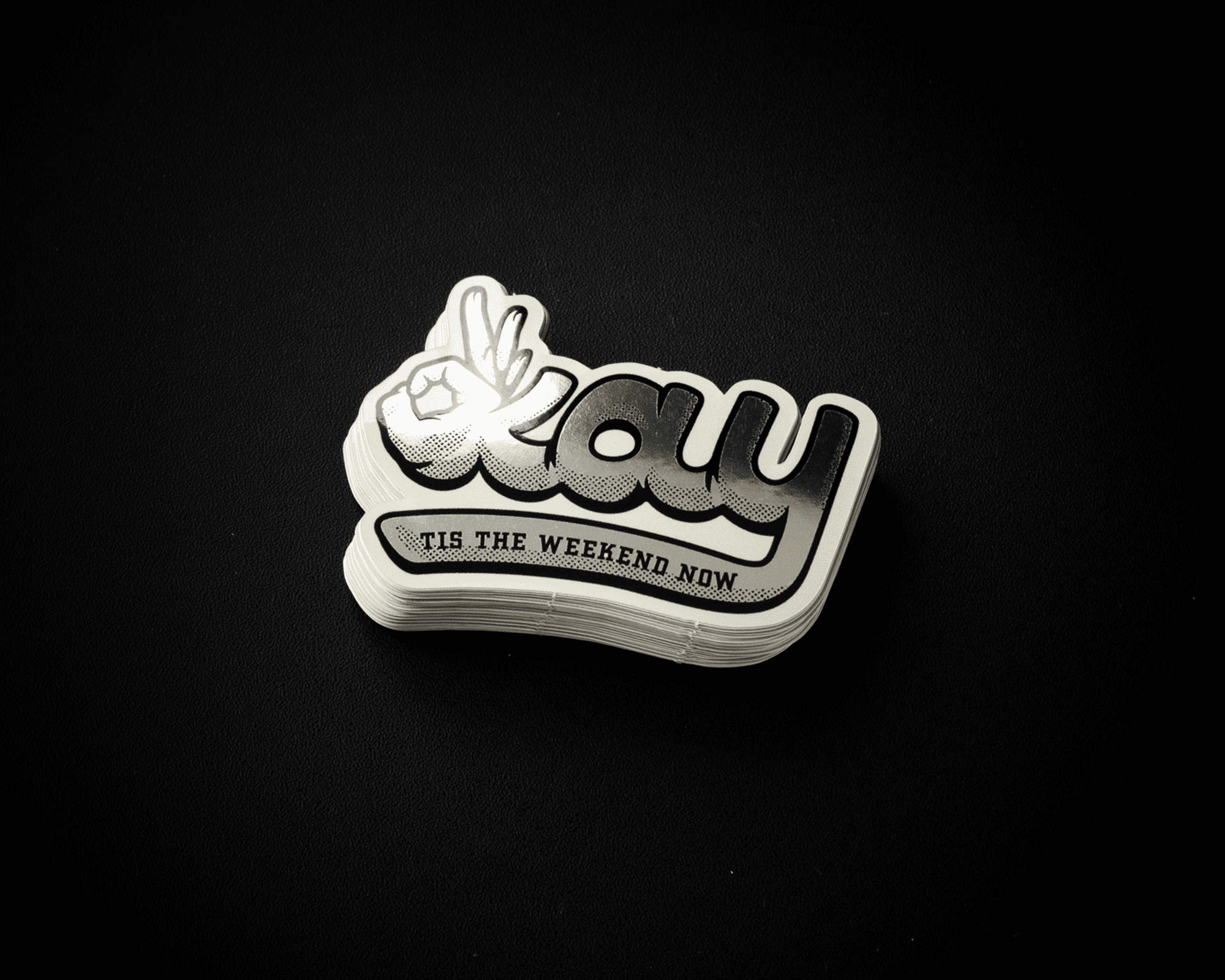 A stack of mirror stickers with the word "okay" and a hand making a peace sign, followed by "Tis the Weekend Now," on a black background.