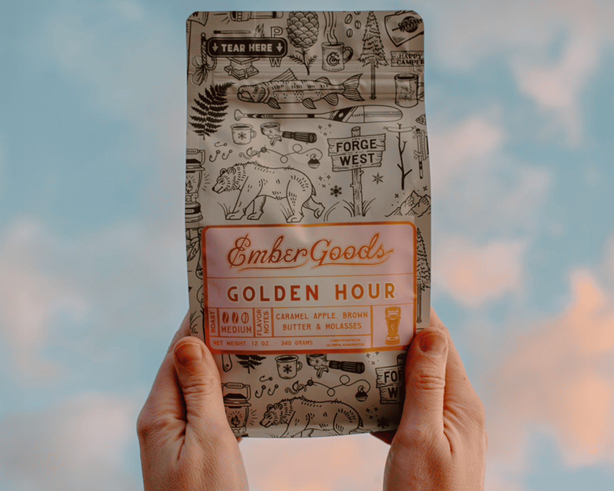 Hände halten eine Tüte Golden Hour Kaffee von Ember Goods mit metallischem Etikett vor Himmelskulisse
