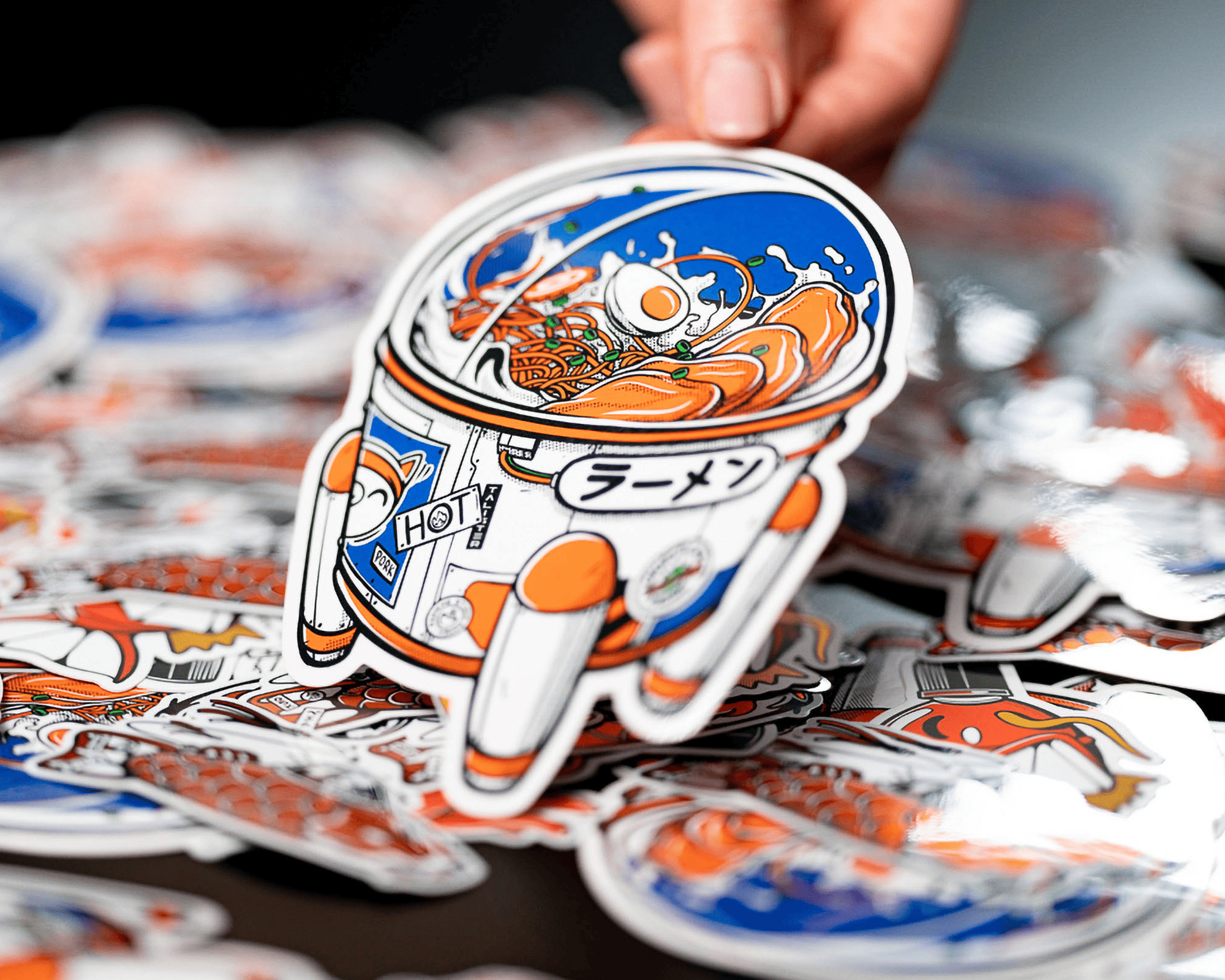 A hand holding a vinyl sticker of an astronaut eating ramen in a space helmet, surrounded by similar stickers.