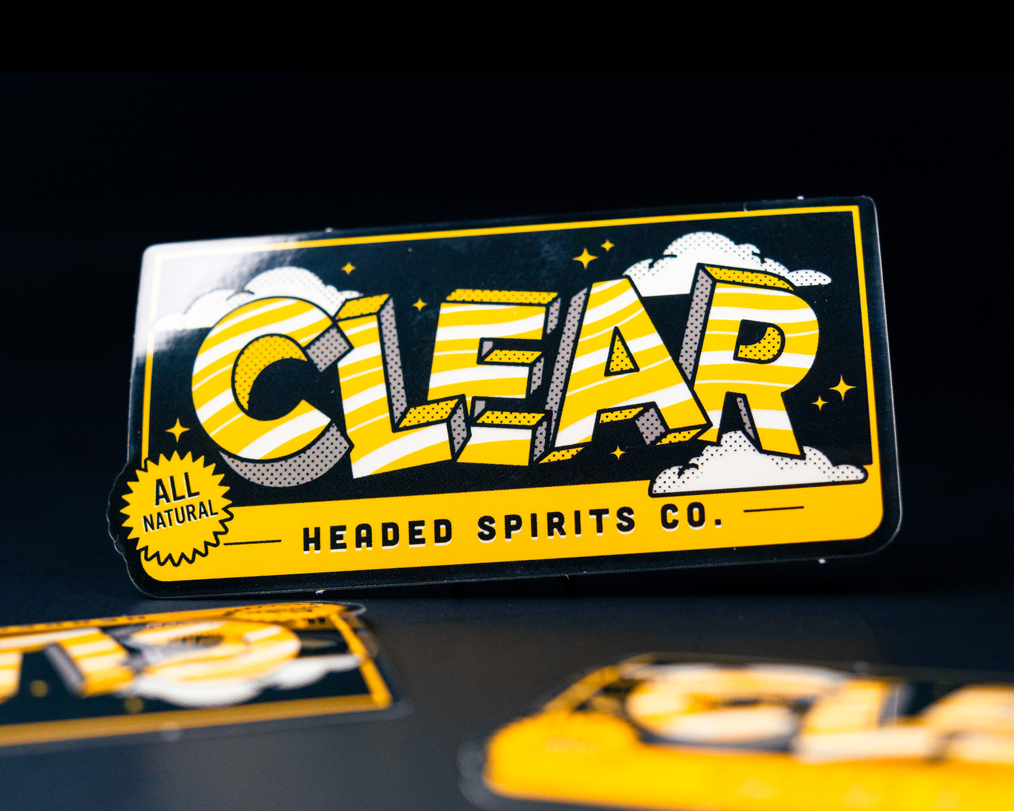 Close-up of a colorful "CLEAR Headed Spirits Co." sticker with a bold design and all-natural label, set against a dark background.