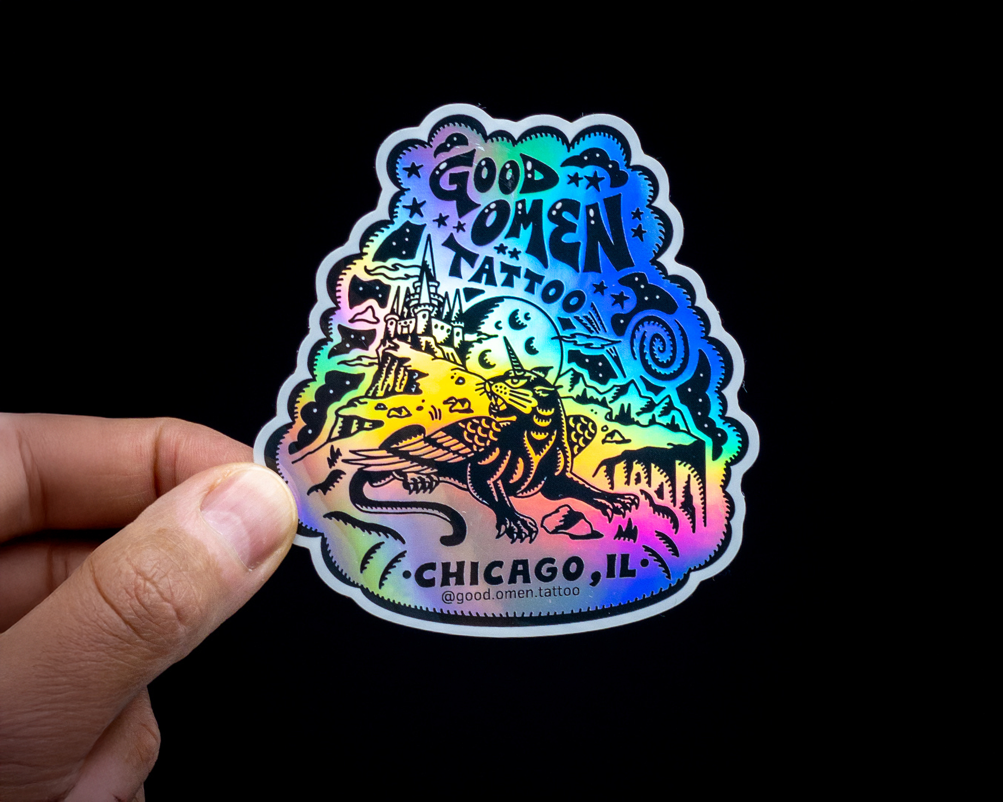 A hand holds a holographic sticker featuring a mythical creature, stars, and a castle, with text "Good Omen Tattoo, Chicago, IL."