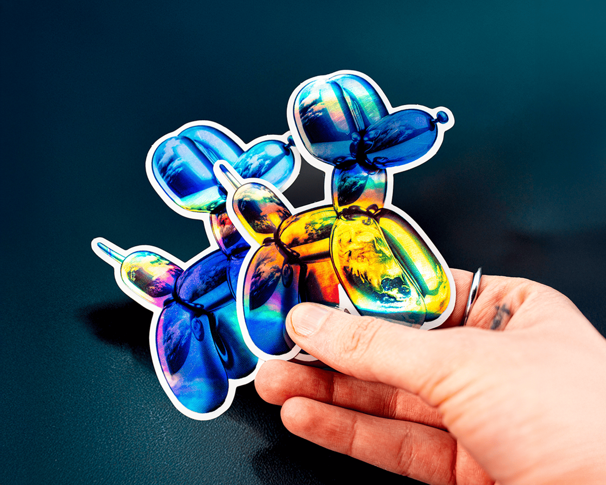 Hand holding holographic balloon dog stickers with a glossy, reflective surface against a dark background.
