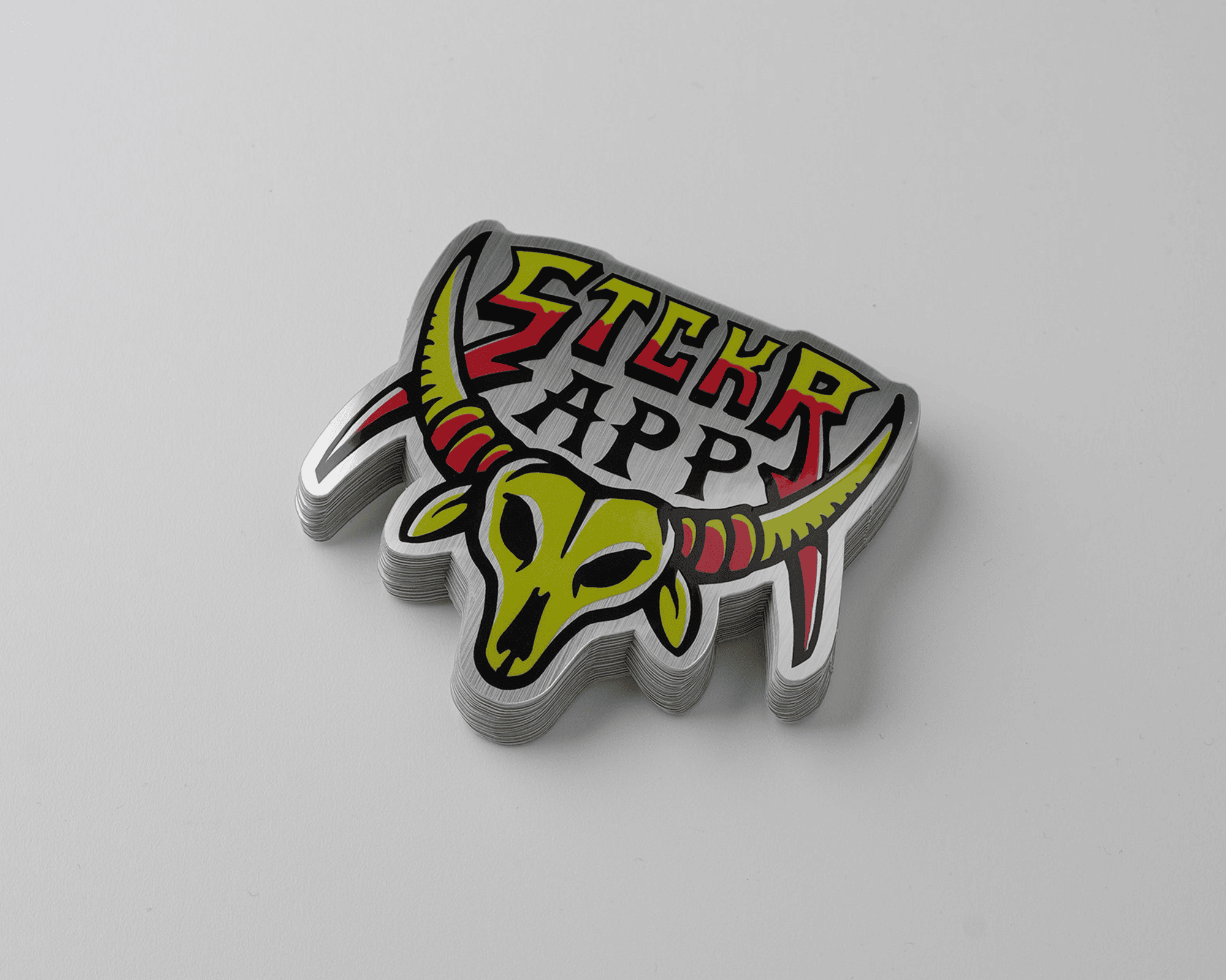 Stack of stickers in brushed aluminum featuring a stylized yellow and red bull skull with large horns and the text "STCKR APP" on a gray background.