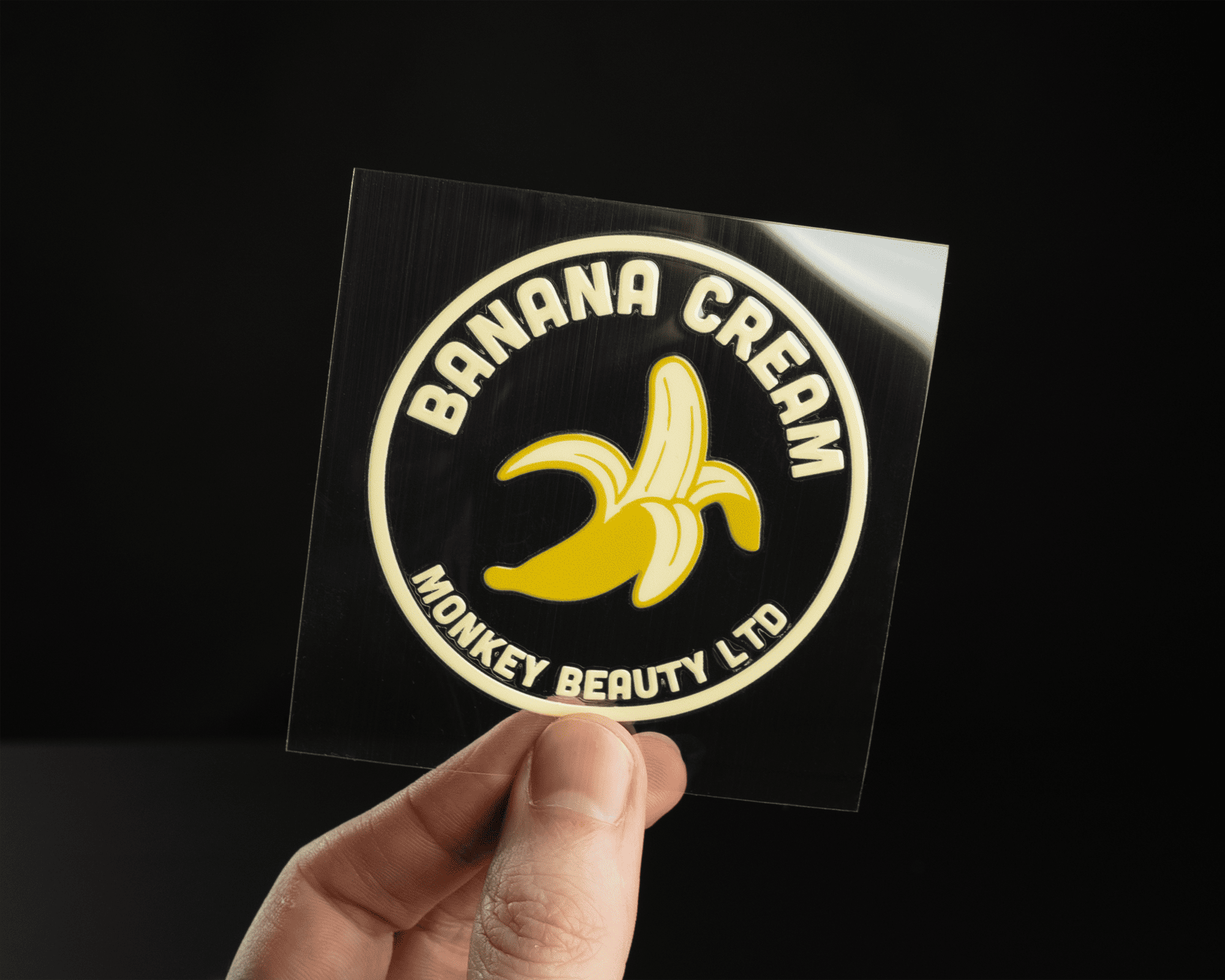 Hand holding a circular Uv dtf sticker with a banana graphic and text: "BANANA CREAM MONKEY BEAUTY LTD" on a black background.