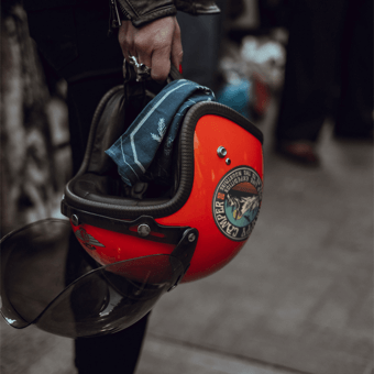 Person holding a red motorcycle helmet with a visor, decorated with a custom sticker, standing on an urban street.