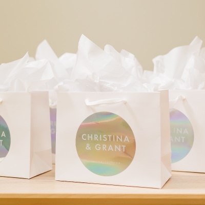 White gift bags with handles placed on a wooden surface, featuring a circular holographic sticker that reads 'CHRISTINA & GRANT' in white text.