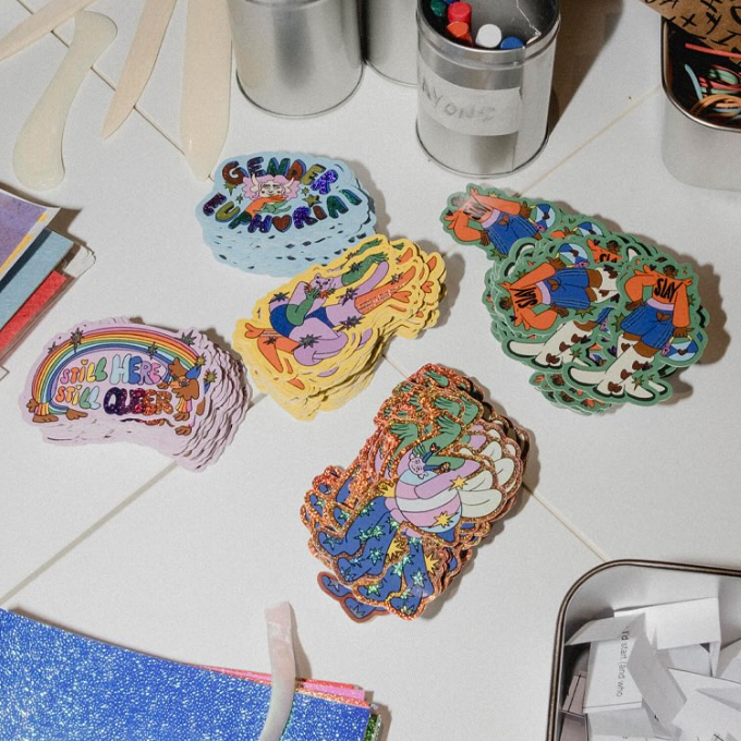 Die-cut stickers in different materials laying on a white table, surrounded by stationary, in a workshop