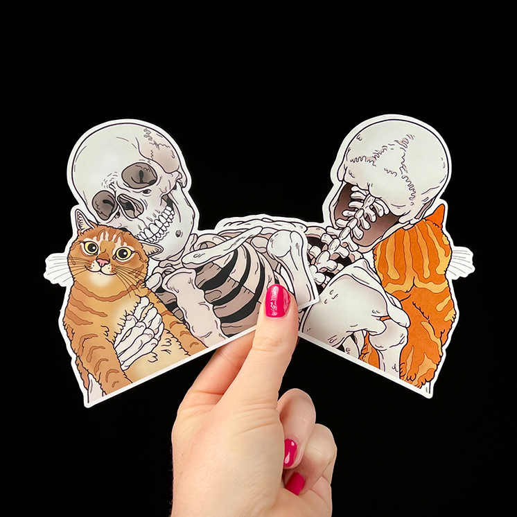 Hand holding a cutout of two skeletons hugging two orange cats against a black background.
