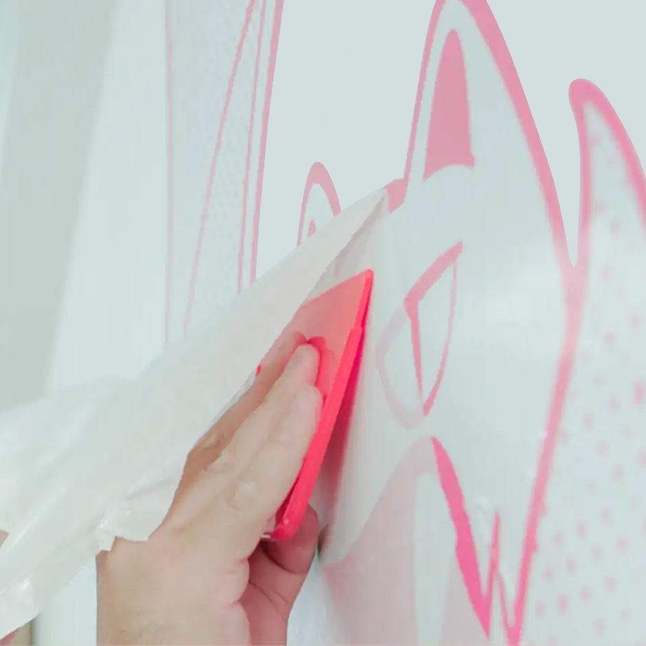 Hand using a red squeegee to apply a pink vinyl transfer of a stylized animal face on a light-colored wall.